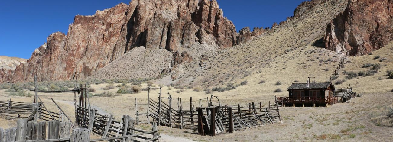 A weathered wooden barn at Birch Creek Historic Ranch stands below a striped, eroded desert peak